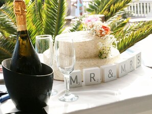 A wedding cake with "M & MRS" blocks, a bottle of champagne, and glasses on a table with palm leaves in the background. - Terezas Hotel (Corfu)