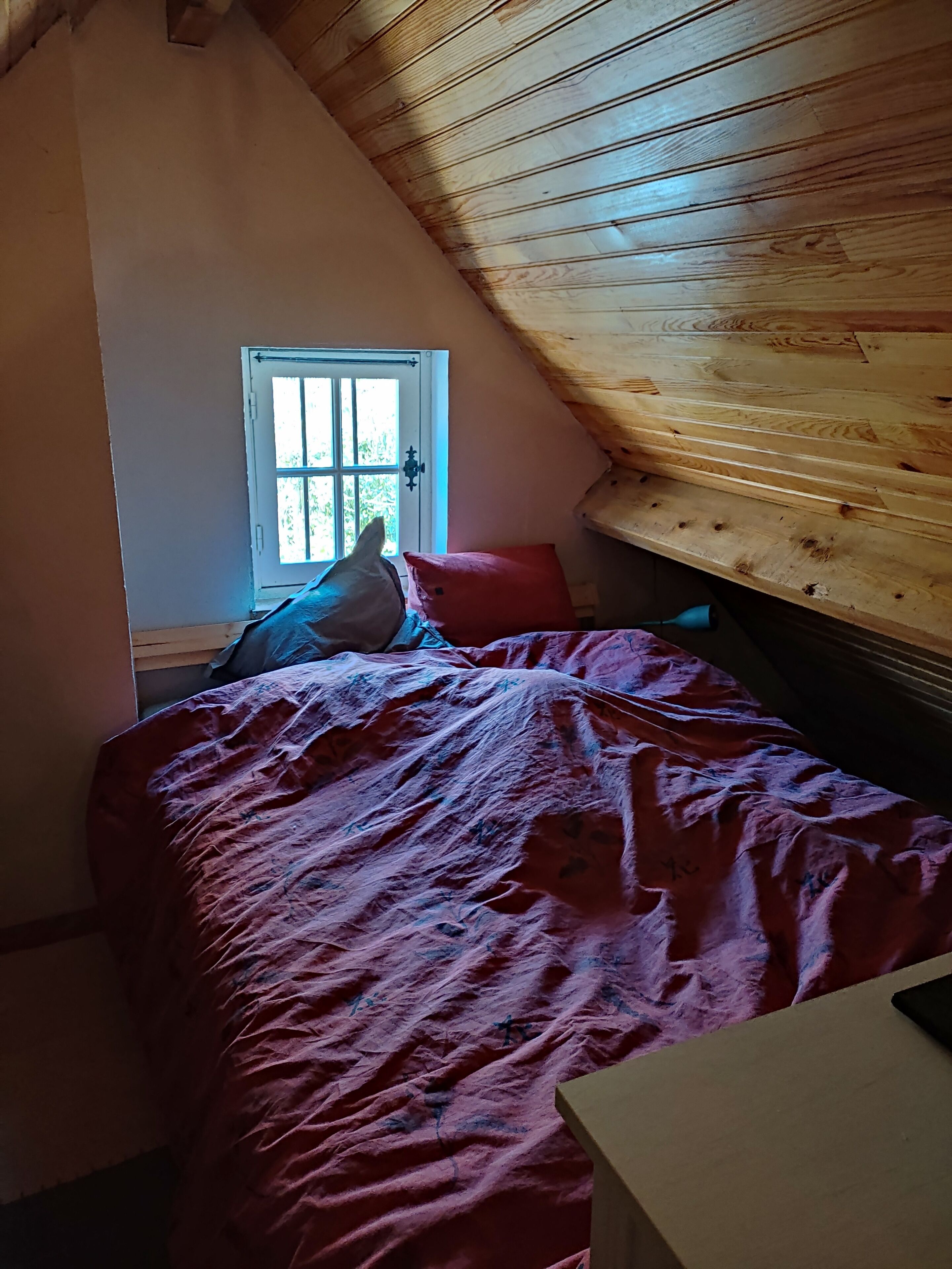 A bedroom with a sloped ceiling, a window with a view of greenery, a bed with a red blanket, and a wooden headboard.