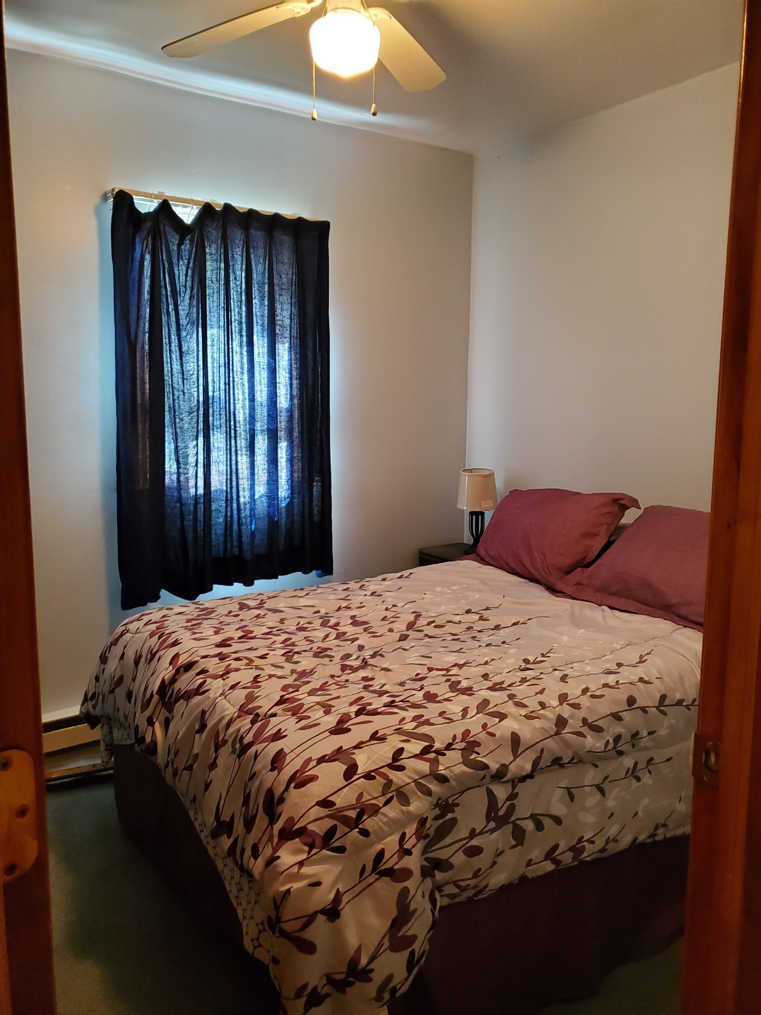 Een slaapkamer met een bed, een plafondventilator, een raam met gordijnen en een nachtkastje met een lamp.