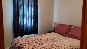 A bedroom with a bed, a ceiling fan, a window with curtains, and a nightstand with a lamp. - Sunset cottage complete w/large deck, fire pit and mins away from public beach (Lake Milton)