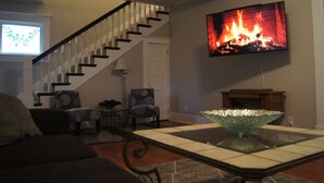 A living room with a staircase, a flat-screen TV displaying a fireplace, and a glass coffee table. - Charming Colonial Home with Private Chipping Green (Hutchinson)