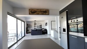 A modern kitchen with a large refrigerator, a dining area with a sofa and chairs, and a view of the outdoors through large windows.