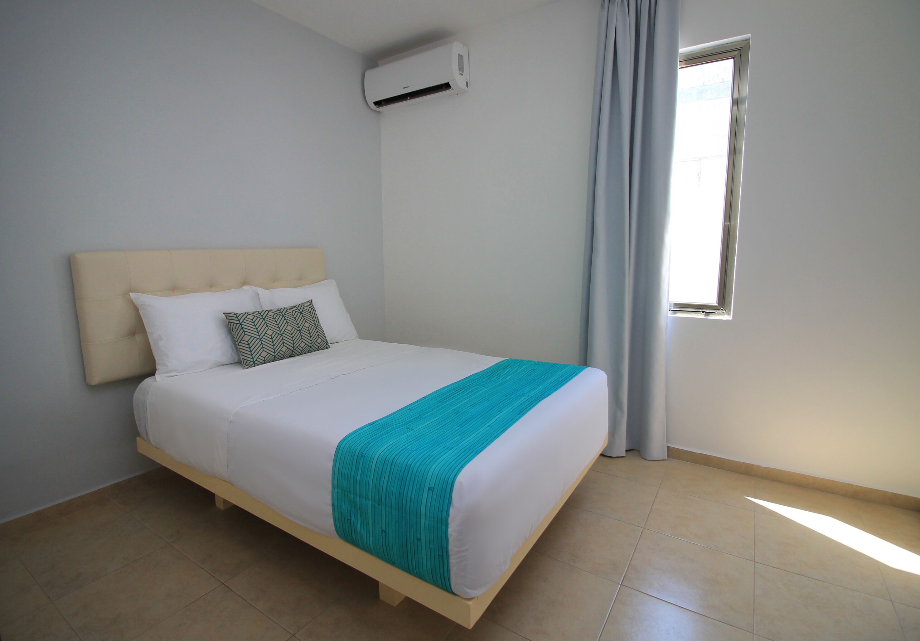 A neatly made bed with a white mattress, a teal blanket, and two pillows. There is a window with curtains, an air conditioning unit on the wall, and a tiled floor.