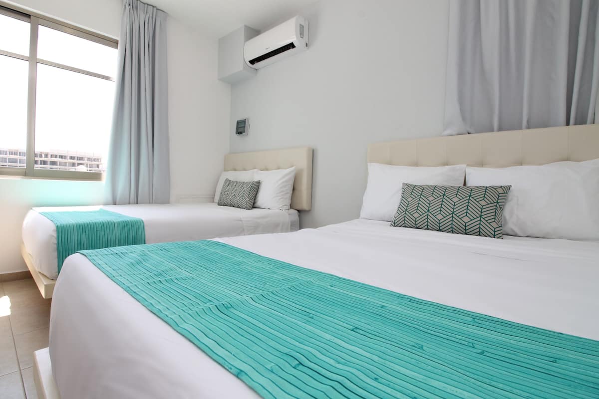 Una habitación de hotel con dos camas, cada una con cabecero blanco y manta color turquesa. Una unidad de aire acondicionado está montada en la pared sobre las camas.