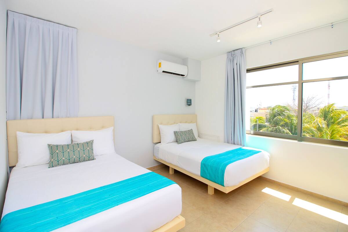 Habitación de hotel con dos camas, ventana con vista a palmeras, aire acondicionado y una lámpara en el techo.
