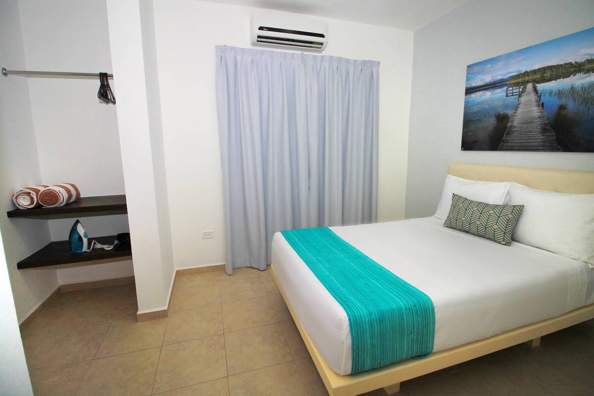 Habitación de hotel con cama, un cuadro de un lago, cortina y estante con toallas y plancha.