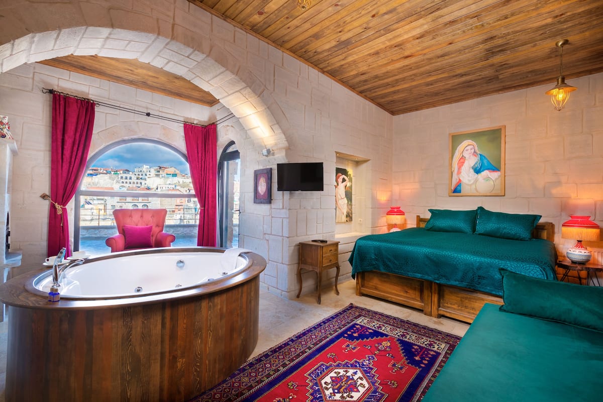 A hotel room with a wooden ceiling, a large bathtub, a bed with green bedding, a red chair, and a view of buildings through an arched window.