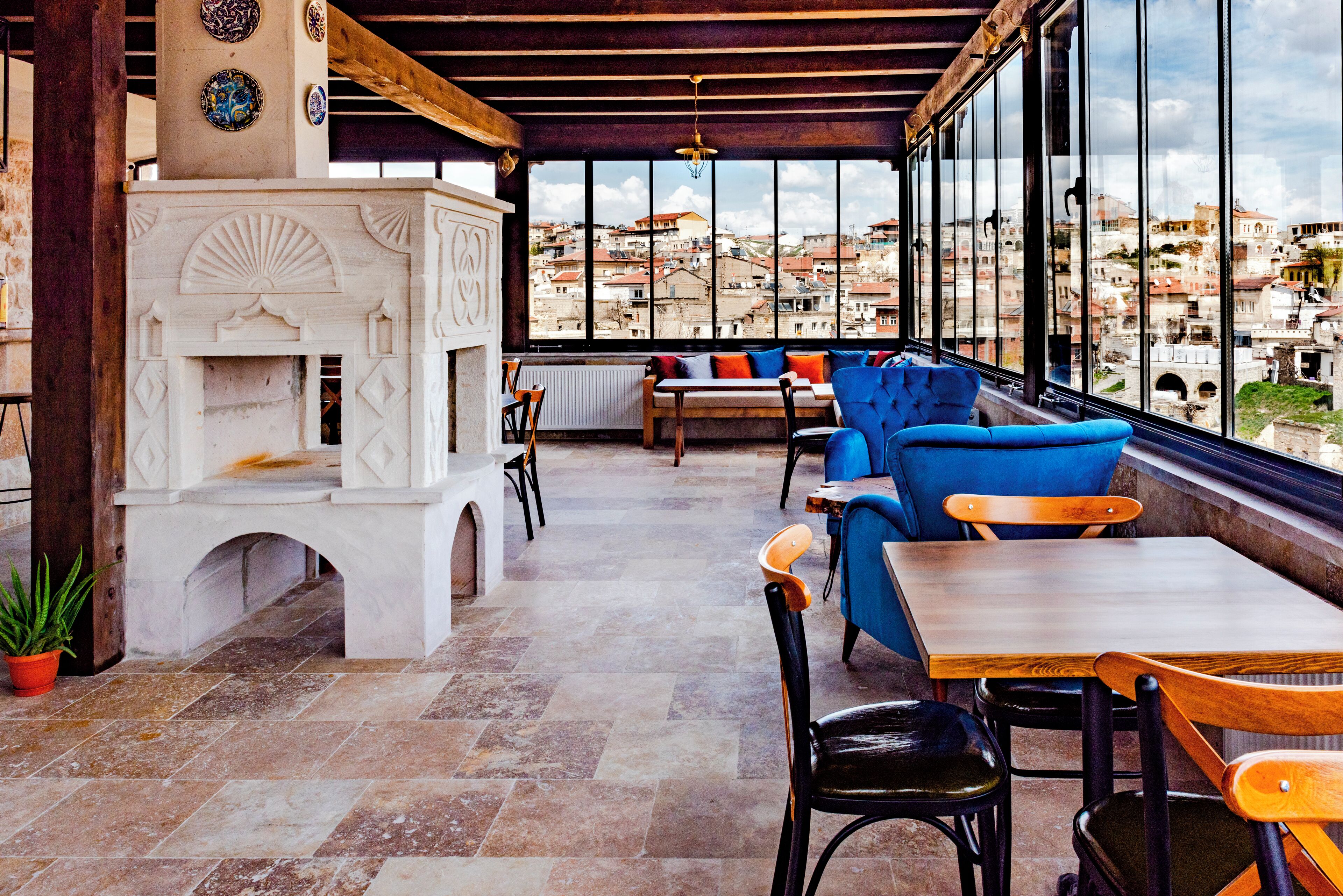 A terrace with stone flooring, a white fireplace, blue and wooden furniture, and a view of a cityscape through large windows.