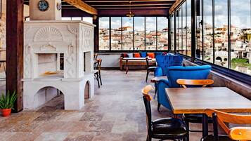 A terrace with stone flooring, a white fireplace, blue and wooden furniture, and a view of a cityscape through large windows.