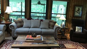 A cozy living room with a sofa, a coffee table with magazines, and a chair. There are two windows with a view of greenery outside.