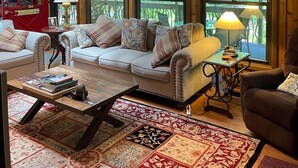 A cozy living room with a large sofa, a coffee table, and a fireplace. There are multiple windows providing natural light and views of the outdoors.
