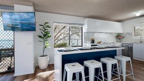 Une cuisine moderne avec des meubles blancs, un îlot central doté de tabourets, une plante en pot et une grande fenêtre donnant sur un pont.