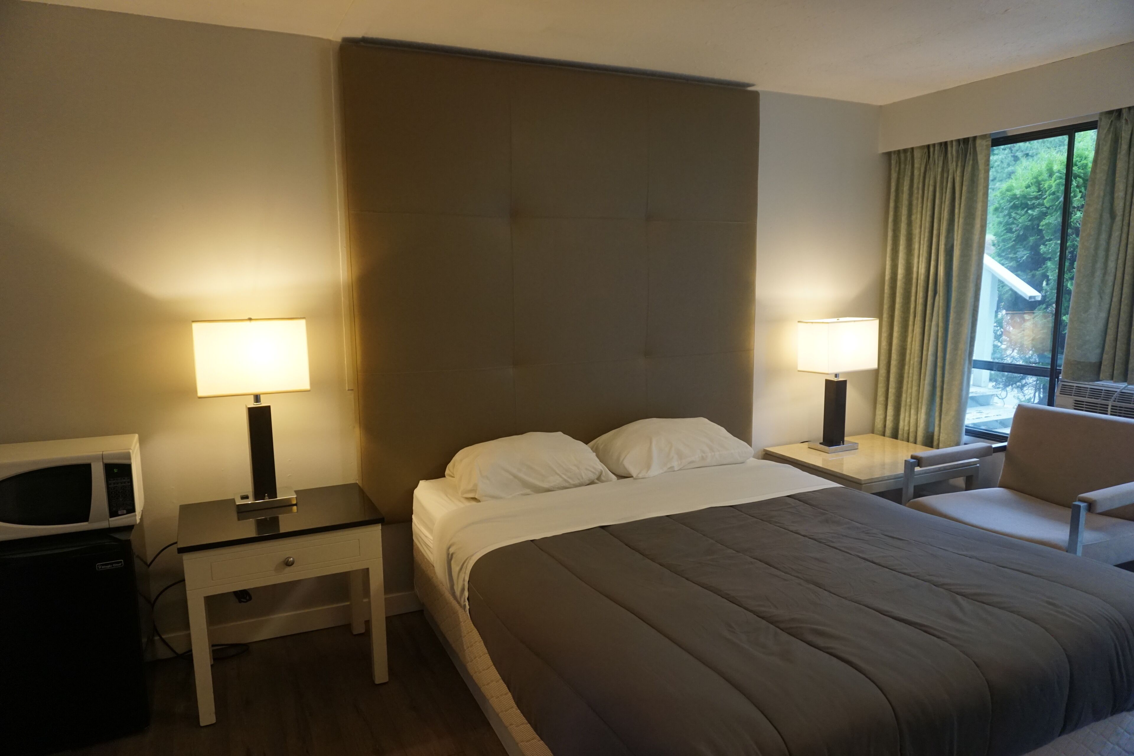 A hotel room with a bed, a desk with a lamp, a chair, a microwave, and a window with curtains.