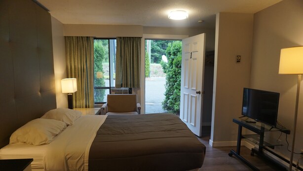 A hotel room with a large bed, a television on a stand, a desk with a lamp, and a view of the outdoors through a door. - The Mighty Fraser Motel (Boston Bar)