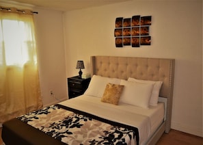 Une chambre à coucher avec une tête de lit capitonnée, un lit recouvert de draps blancs, un plaid à motifs, une table de chevet avec une lampe et une œuvre d’art murale au-dessus du lit.