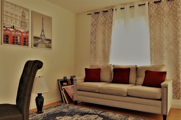 Un salon avec un canapé beige, des coussins rouges, un fauteuil foncé, un tapis à motifs et deux tableaux encadrés accrochés au mur.