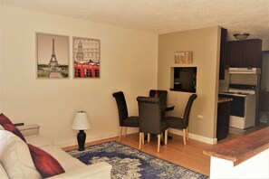 A compact living space with a dining area, a sofa, a lamp, and a kitchen area. - Urbana – Charming, Bright 2-Bedroom, 1.5-Bath Apartment in the Heart of Grand Forks (Grand Forks)