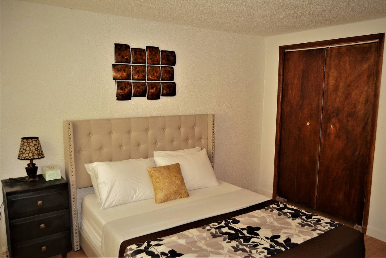 A bedroom with a tufted headboard, a bed with white linens and a patterned throw, a nightstand with a lamp, and a wooden wardrobe.