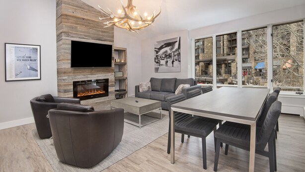 A modern living room with a fireplace, a flat-screen TV, comfortable seating, and large windows offering a view of the city. - Unit 14 WITH AC-NEW LOWER PRICE. HEART OF VAIL. WALK TO GONDOLA (Vail)
