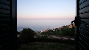 Une vue sur la côte à travers des volets ouverts, avec une route, de la végétation et des bâtiments visibles au loin.