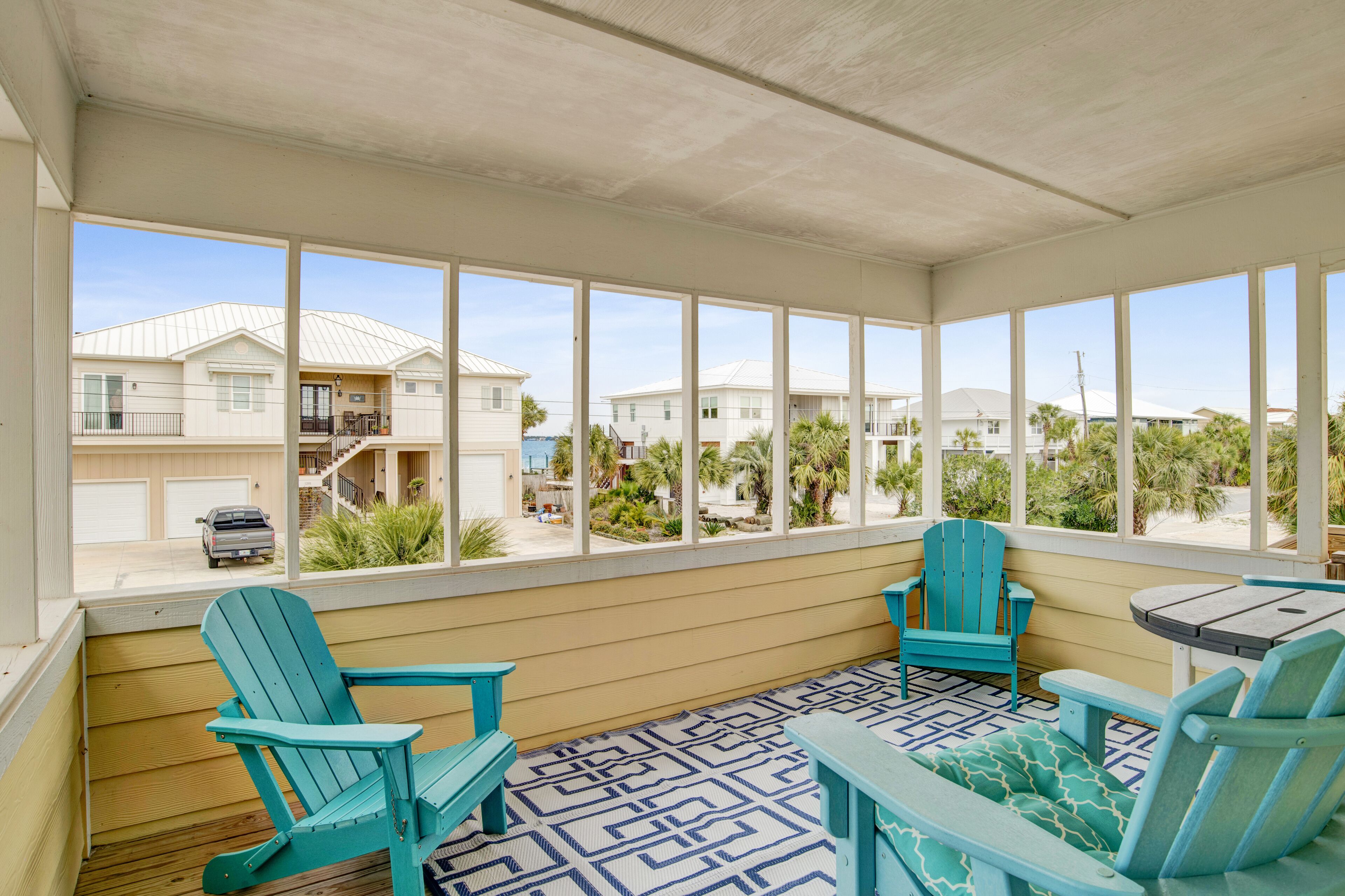 Un espace extérieur abrité, avec vue sur un quartier résidentiel, comprenant une terrasse en bois, des fauteuils Adirondack turquoise et une petite table.