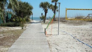 Un chemin en béton mène à une plage de sable bordée de palmiers et surmontée d’un poteau de but.