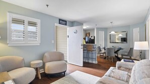 Un salon avec un canapé, un fauteuil et une petite table d’appoint. On aperçoit à l’arrière-plan un coin repas avec des chaises et une table.