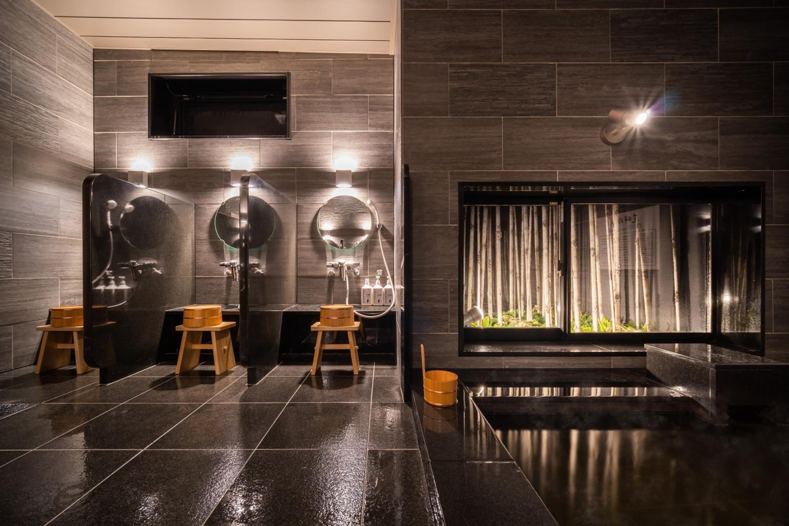 A modern bathroom with dark tiled floors, wooden stools, and a large mirror.