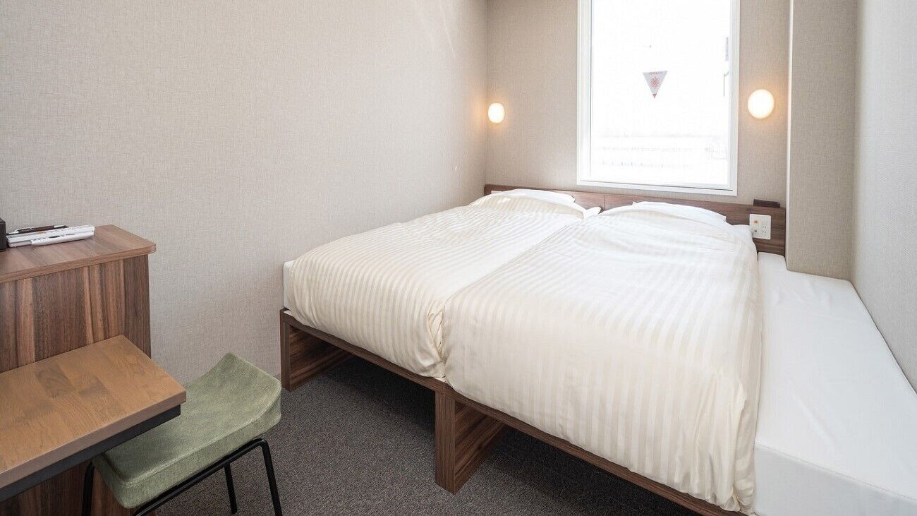 A single bed with a wooden frame, a small desk with a chair, and a window with a view of a building.