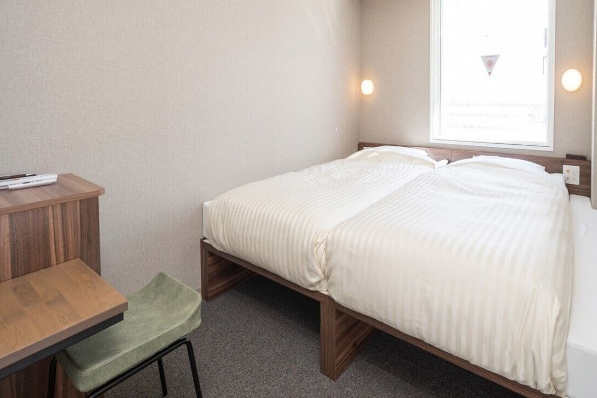 Una cama individual con estructura de madera, un escritorio pequeño con una silla y una ventana que da a un edificio.