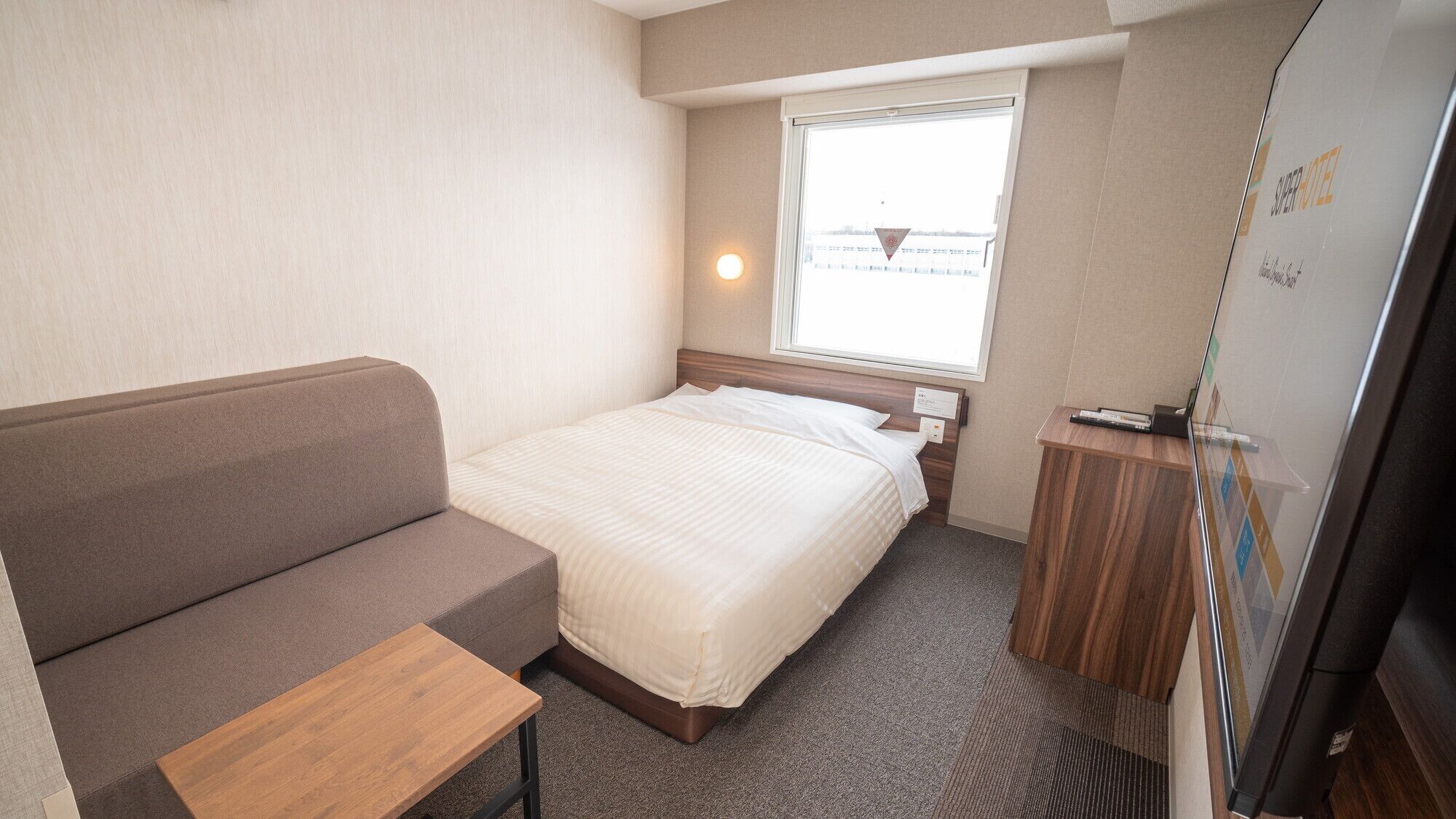 A bedroom with a bed, a sofa, a small table, a window, and a wall-mounted TV.