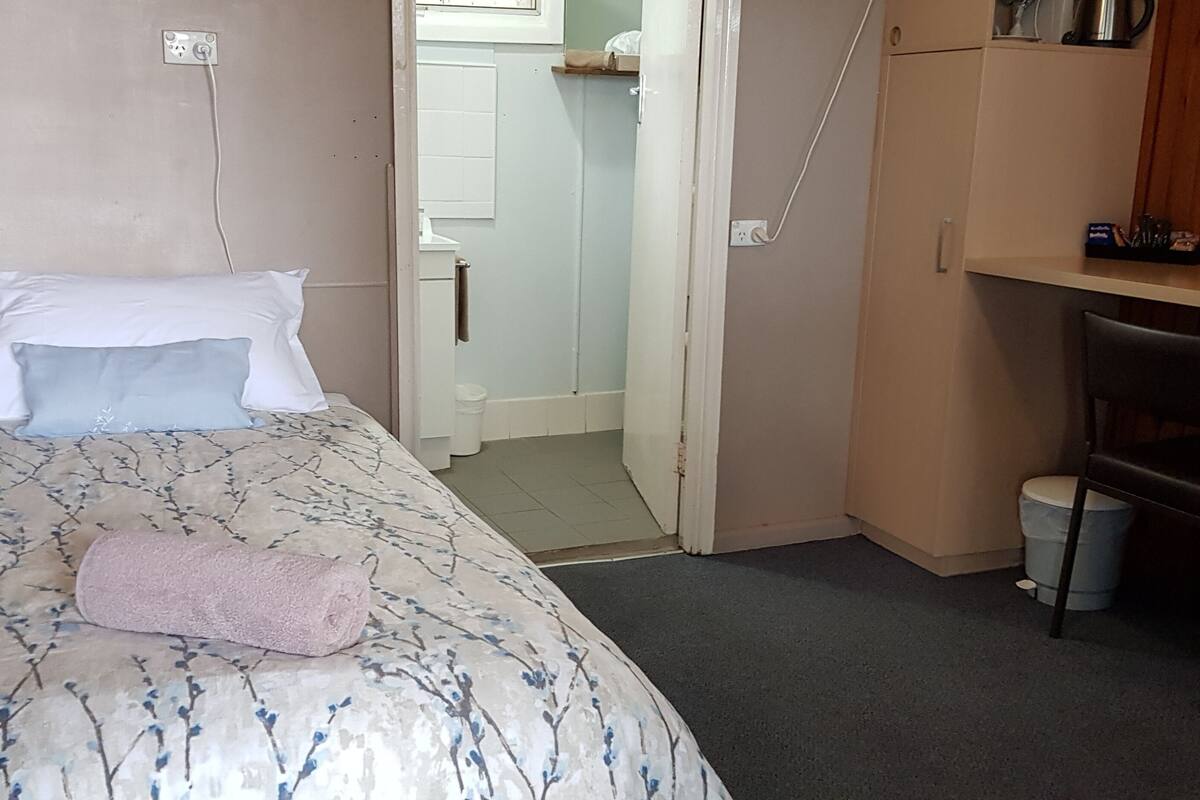 Una habitación pequeña y funcional con cama, escritorio y silla. Hay una puerta que da a un baño.
