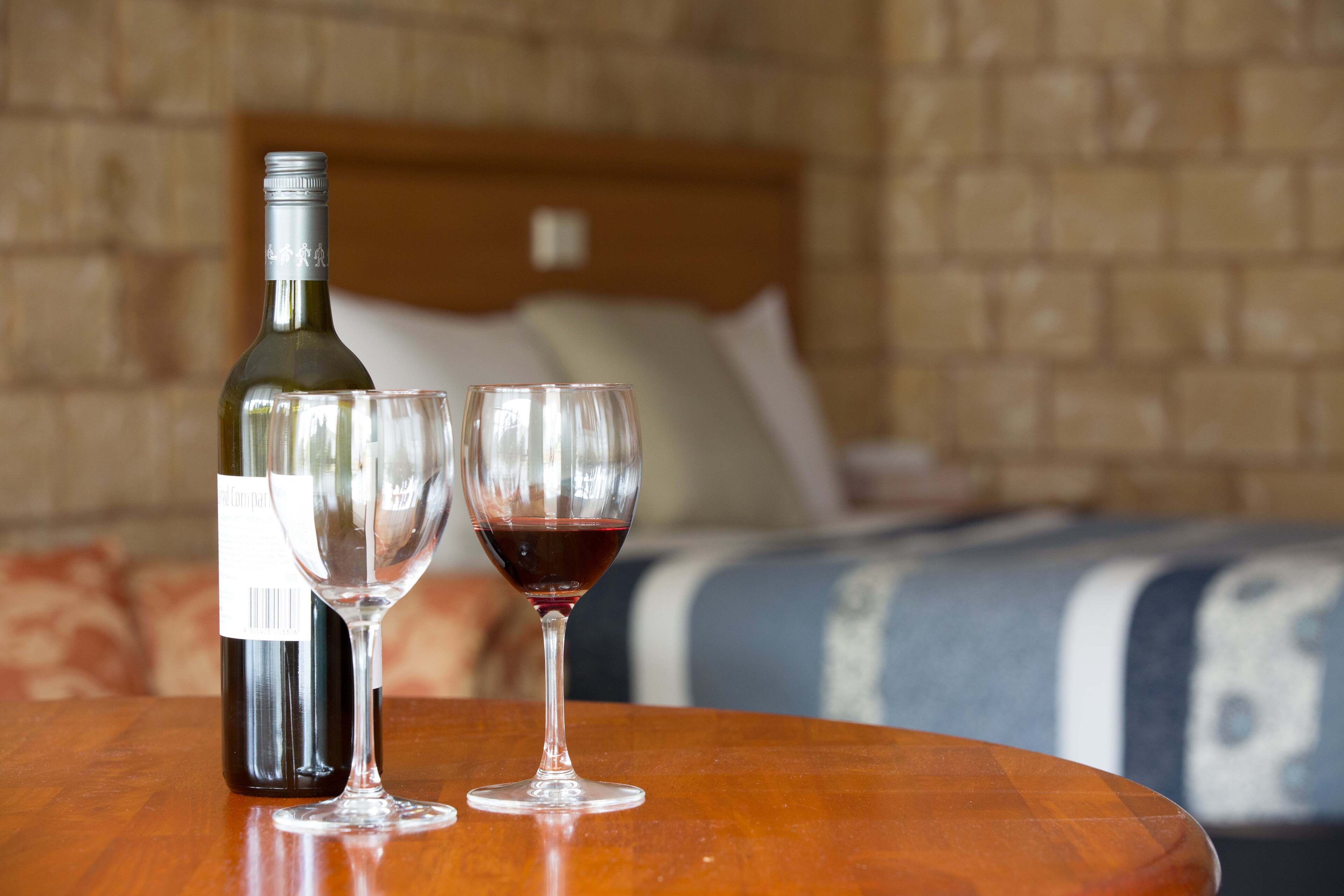A bottle of wine and two wine glasses on a wooden table in a hotel room with a brick wall and a bed in the background.