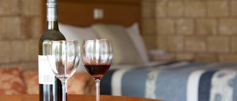 Una botella de vino y dos copas sobre una mesa de madera en una habitación de hotel, con una pared de ladrillo y una cama al fondo.