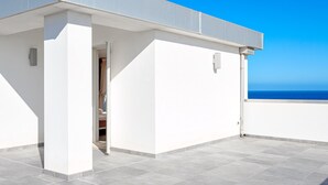 A rooftop terrace with a clear view of the sea, a white building, and a door leading to an interior space.