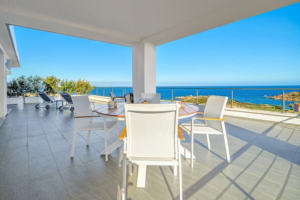 Une terrasse avec une table et des chaises, une vue sur la mer et un bâtiment blanc en arrière-plan.