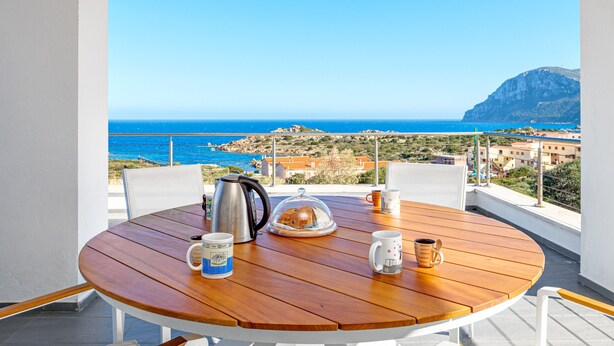 A round wooden table with a kettle, a glass dome, and two mugs on a balcony overlooking a coastal view.