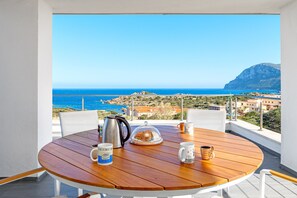 A round wooden table with a kettle, a glass dome, and two mugs on a balcony overlooking a coastal view. - Penthouse Apartment 12 in Villa Marconi with Sea & Mountain View and Panoramic Terrace (Golfo Aranci)