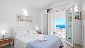 A bedroom with a bed, bedside tables, a painting, and a view of the sea through an open door.