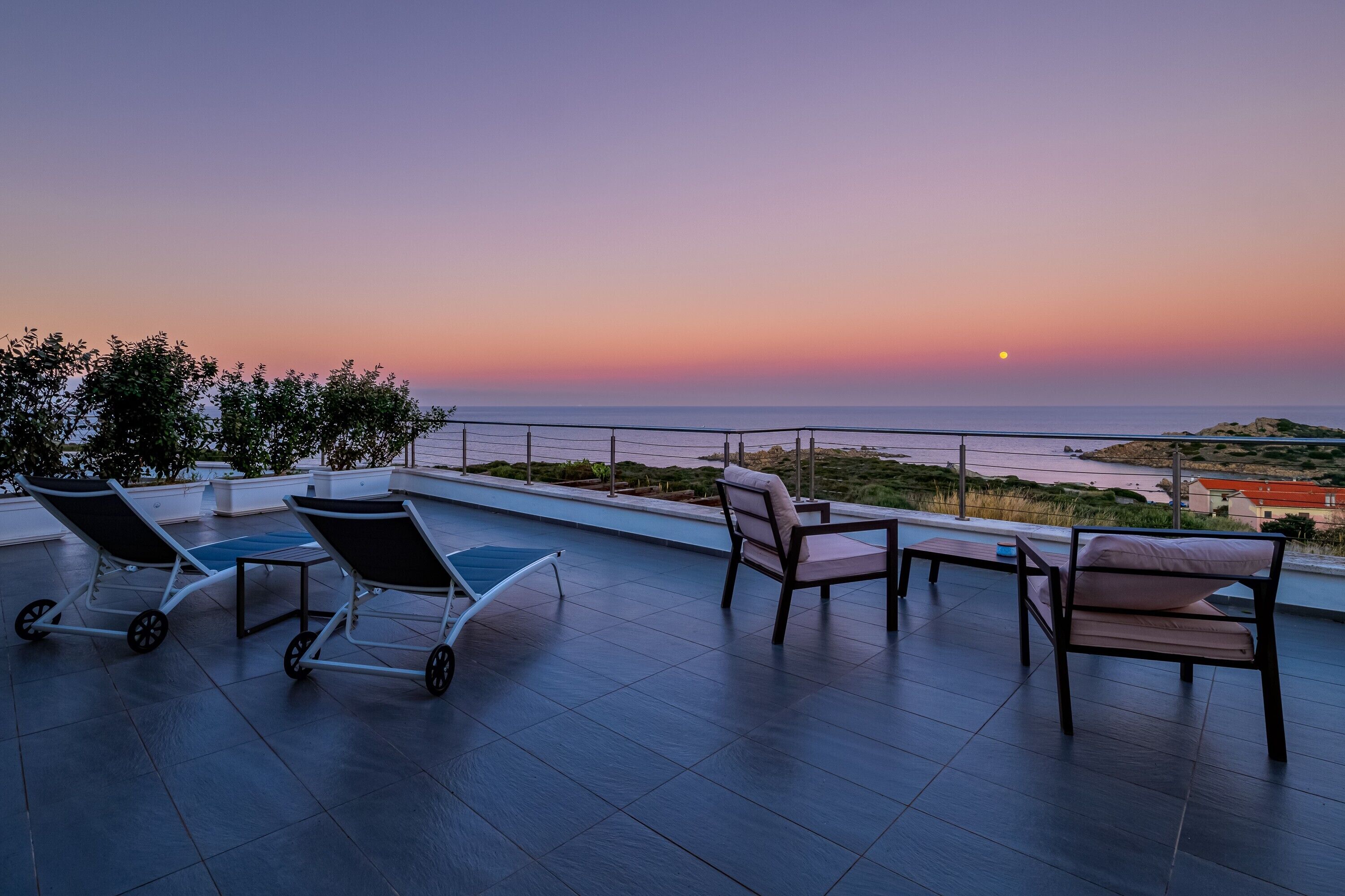 Une terrasse avec des fauteuils et une table, d’où l’on peut admirer un paysage côtier au coucher du soleil.