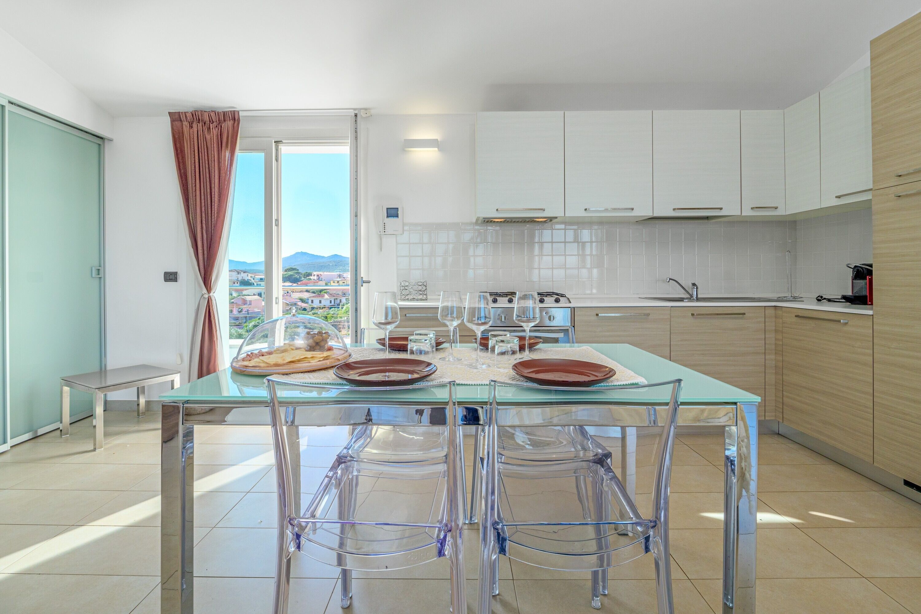 Una cocina moderna con una mesa de comedor de vidrio, sillas transparentes, armarios blancos y vistas de la ciudad.