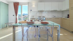 A modern kitchen with a glass dining table, clear chairs, white cabinets, and a view of the city.