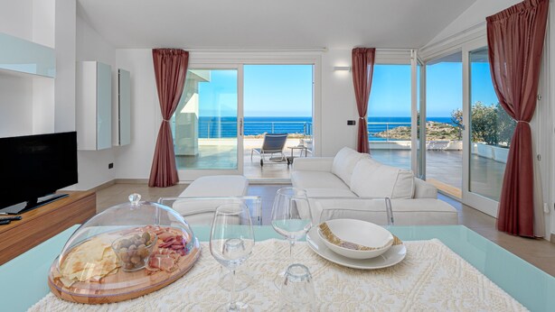 Un salon moderne doté d’un téléviseur, d’une table à manger avec de la verrerie, et offrant une vue sur la mer grâce à de grandes fenêtres.