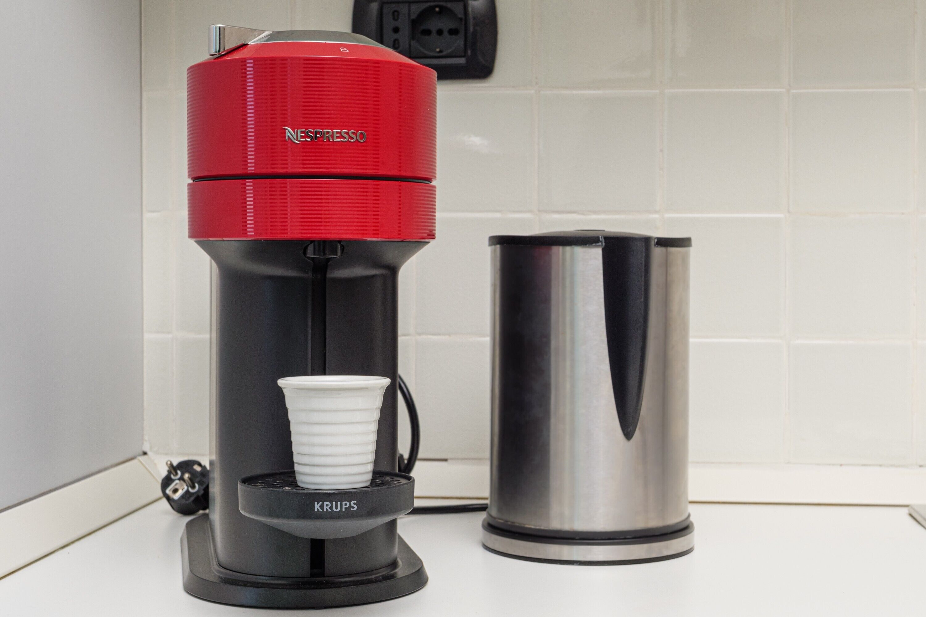 Nespresso-kahvinkeitin, jossa on Krupsin kahvikapselikotelo, ruostumattomasta teräksestä valmistettu kahvipannu ja taustalla näkyvä pistorasia.