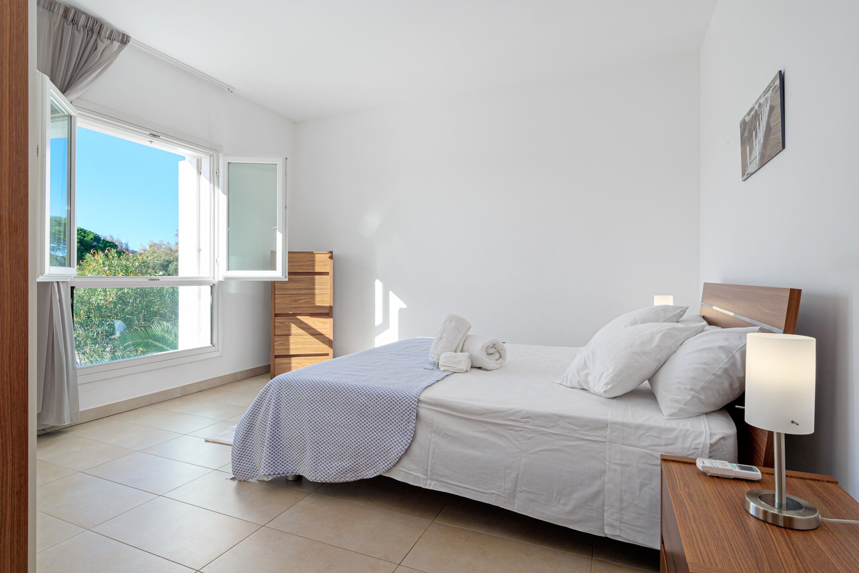 Une chambre à coucher avec un lit, une table de chevet avec une lampe, une fenêtre avec des rideaux et une vue sur la verdure à l’extérieur.