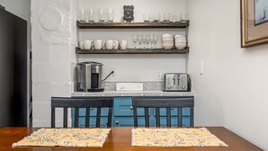 A dining area with a wooden table and chairs, a coffee machine, a toaster, and shelves with glassware and cups.