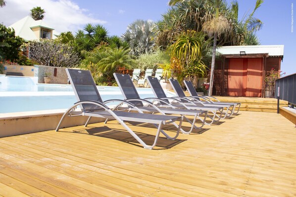 Una terraza de madera con tumbonas mirando una piscina, rodeada de plantas tropicales y un edificio con una puerta roja.