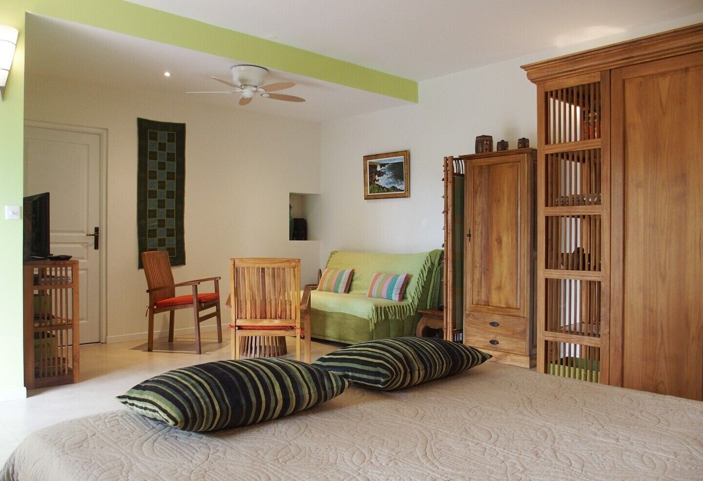 A bedroom with a bed, two chairs, a green sofa, a wooden wardrobe, a ceiling fan, and a framed picture on the wall.