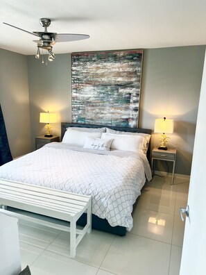 A bedroom with a bed, two bedside tables with lamps, a ceiling fan, and a large abstract painting on the wall.