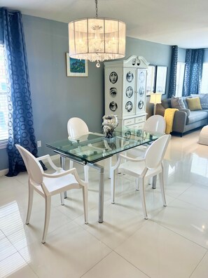 A dining area with a glass table and white chairs, a chandelier, a cabinet with decorative plates, and a sofa with cushions.